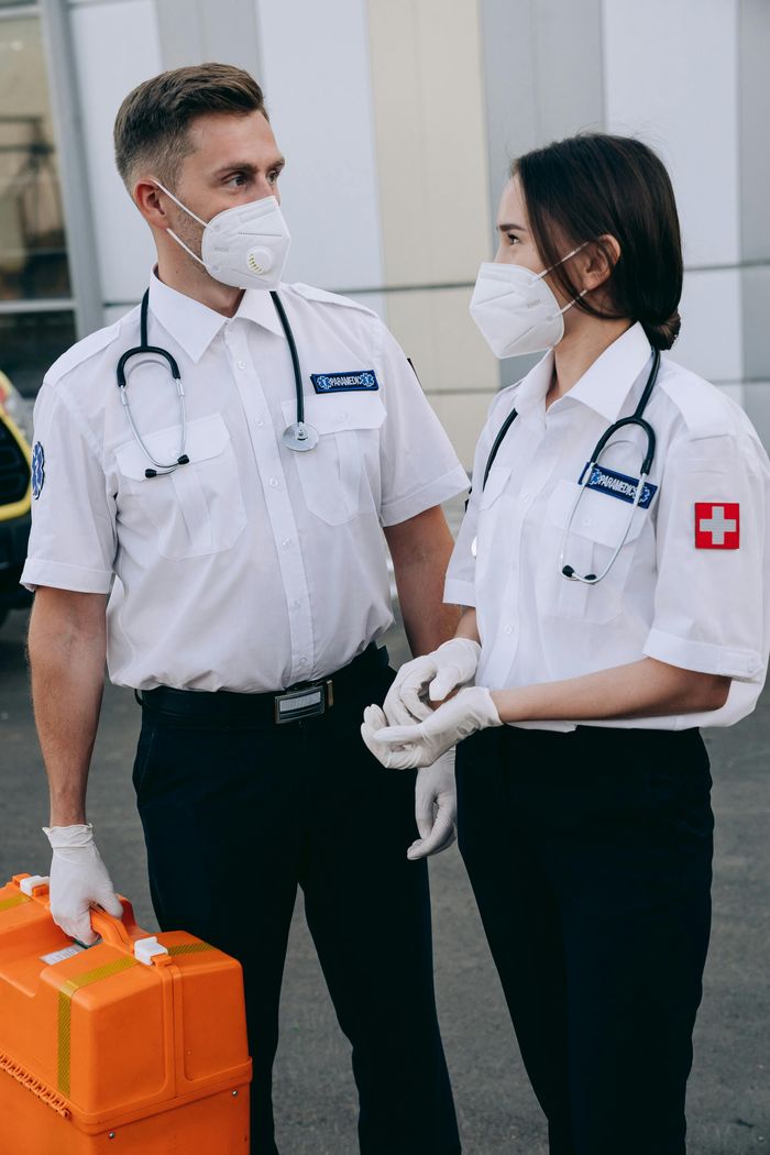 Two paramedics wearing masks and stethoscopes, standing outdoors with emergency medical kit.