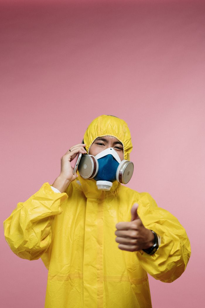 Person in yellow suit wearing a respirator on a pink background giving a thumbs up.